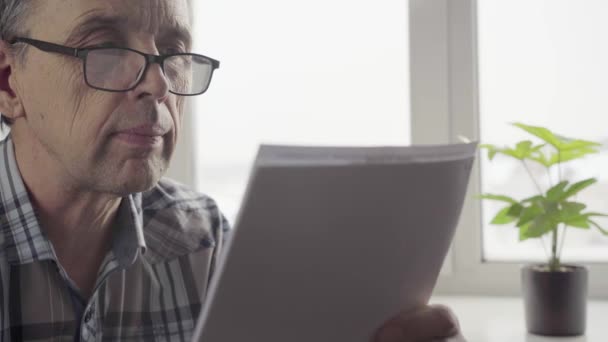 Um homem idoso com óculos lê atentamente o documento nos lençóis — Vídeo de Stock