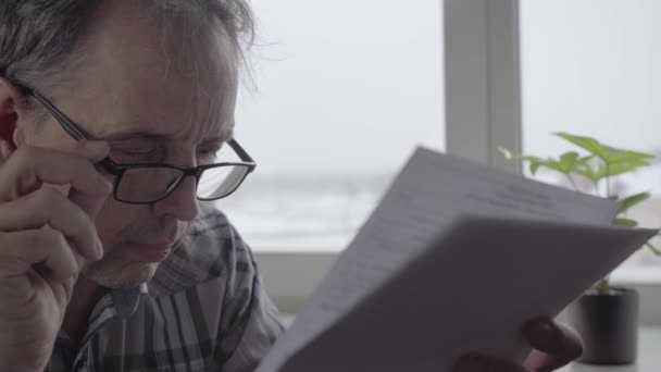 Ένας ηλικιωμένος άνδρας με γυαλιά διαβάζει προσεκτικά το έγγραφο στα φύλλα — Αρχείο Βίντεο
