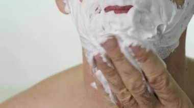 Yüzüne köpük sür. Yaşlı bir adamın yüzüne yakın plan, beyaz arka planda çenesine köpük sürmeye odaklanmış.