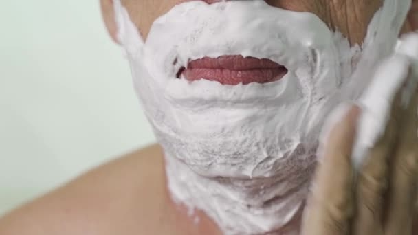 Aplica la espuma en la cara. Primer plano de una cara de hombre mayor, concentrándose en aplicar espuma a su barbilla sobre un fondo blanco — Vídeos de Stock