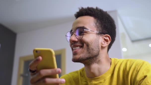 若い、幸せなアフリカ人男性は彼のスマートフォン上で笑顔とテキスト。自宅でスマートフォンを使って幸せな黒人男性 — ストック動画
