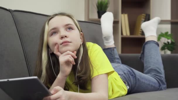 Een gelukkig duizendjarig blank tienermeisje liggend op een comfortabele bank luisterend naar muziek op een tablet — Stockvideo