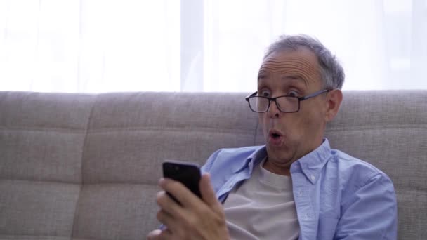 Ο ηλικιωμένος άνδρας είναι σε μεγάλο βαθμό έκπληκτος, κοιτάζοντας την οθόνη smartphone — Αρχείο Βίντεο