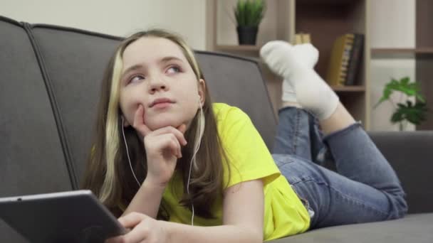 En glad tusenårig vit tonårstjej som ligger på en bekväm soffa och lyssnar på musik på en surfplatta — Stockvideo