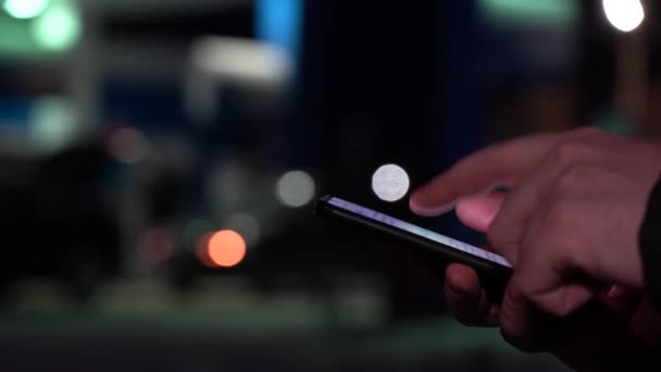 Hand eines Mannes mit Smartphone in der Nacht in der Stadt vor dem Hintergrund verschwommener Lichter von Autos — Stockvideo