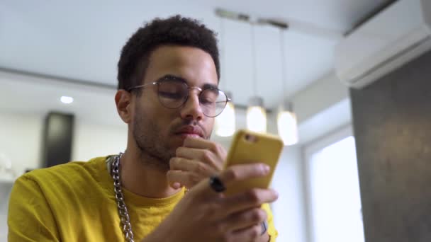 A young man wearing glasses reads messages on a smartphone at home — Αρχείο Βίντεο