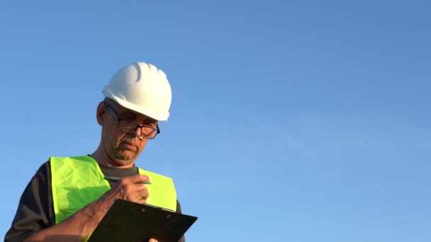 Un anciano con gafas y un casco de trabajo toma notas contra un cielo azul claro — Vídeo de stock