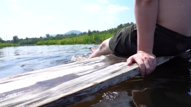 Ein Mann genießt seinen Sommerurlaub, indem er mit den Füßen auf einem Holzbrett im Fluss sitzt — Stockvideo