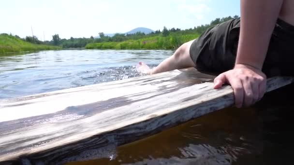 En man njuter av sin sommarsemester genom att sitta på en träskiva med fötterna i floden — Stockvideo