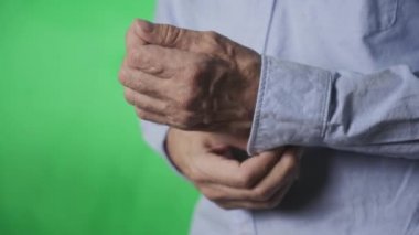 Gömleğinin kolundaki düğmeyi ilikleyen yaşlı bir adamın elleri, yeşil bir arka planda.