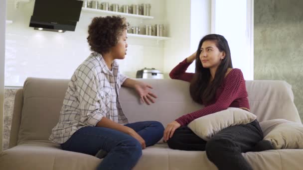 Cámara lenta: Dos amigas, una afroamericana y una asiática, charlando entre ellas mientras están sentadas en casa en el sofá — Vídeo de stock