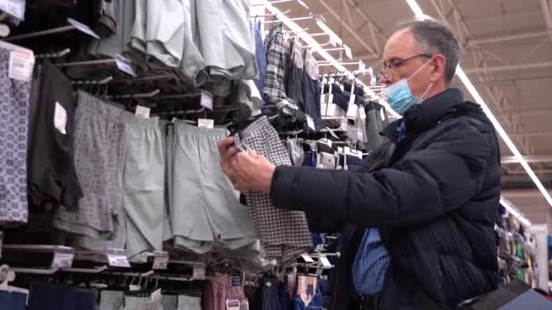 Un anciano elige ropa en una tienda. Un anciano con una máscara debido al coronavirus elige pantalones cortos para dormir en una tienda de ropa — Vídeos de Stock