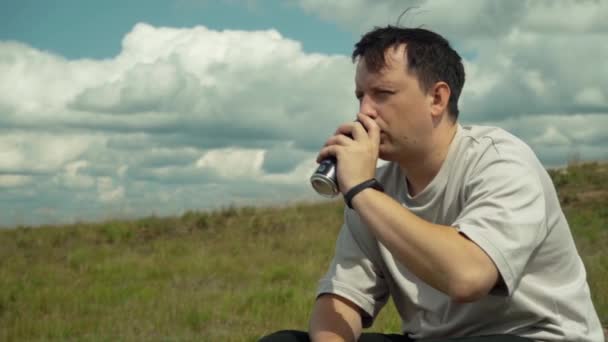 Молодий чоловік сидить на пагорбі і п'є напій з алюмінію на тлі хмар — стокове відео