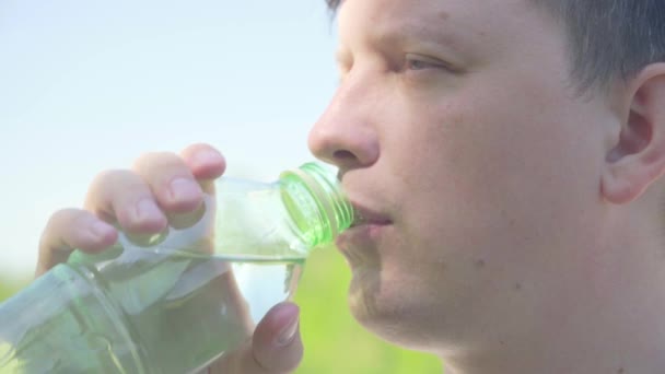 Sete estinguente. Beva l'acqua pulita da una bottiglia. Un giovane di aspetto caucasico beve acqua da una bottiglia di plastica trasparente contro un cielo blu con nuvole bianche. Primo piano — Video Stock