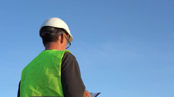 Un anciano con gafas y un casco de trabajo toma notas contra un cielo azul claro — Vídeo de stock