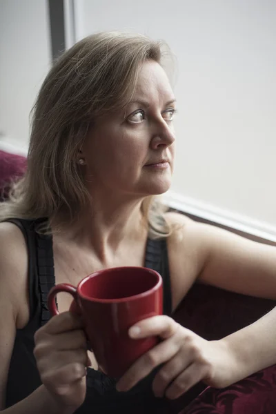 Blond kvinna med röd kaffekopp som sitter vid ett fönster — Stockfoto