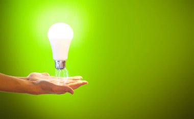 El ele tutuşan ampul. Eko-enerji kavramı, yenilik. Kadın elleri yeşil arka planda elektrik yüklü bir LED ampulü tutuyor..