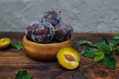 Olgun, sulu erikler, ahşap mutfak masasında yarıya bölünmüş ve gri duvara yaslanmış yeşil yapraklar ve dallar, hasat konsepti, yemek pişirme tatlısı, salata, turta tarifi, kuru erik.