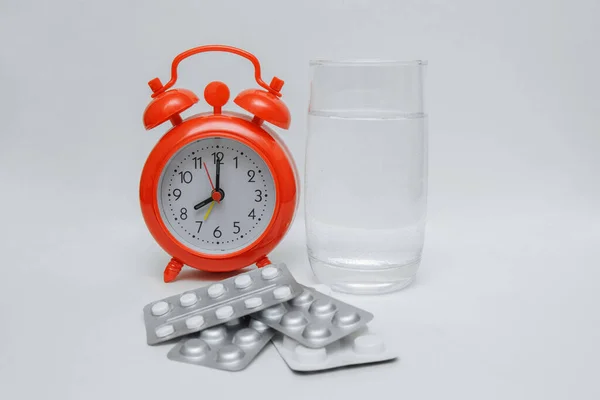 Beyaz arka planda bir bardak su ve haplı kırmızı alarm saati. Metin, sağlık konseptleri, ilaç rejimleri, tedavi, vitaminler ve antibiyotikler için boş bir alan var.