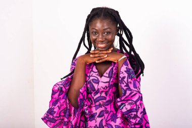 Beyaz arka planda duran, elleri çenesinin altında gülümseyen kameraya bakan Afrikalı genç bir kadın..