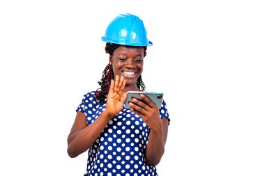 Genç bayan mühendis mavi güvenlik kaskı takıyor ve gülümserken cep telefonuyla yaptığı video görüşmesi sırasında elini sallıyor..