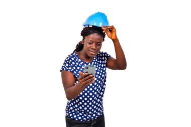 Güvenlik kaskı takmış güzel bir kadın mühendis beyaz arka planda duruyor ve şaşırmış bir şekilde cep telefonuna bakıyor..