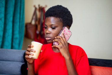 Kırmızı tişörtlü genç kız evde bir kanepede oturmuş elinde tek kullanımlık bir bardakla cep telefonuyla konuşuyor..