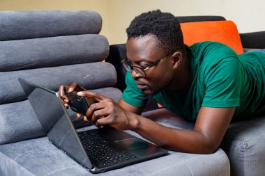 Optik gözlüklü genç adam evdeki kanepede uzanıyor ve cep telefonuyla dizüstü bilgisayarın önünde fotoğraf çekiyor..