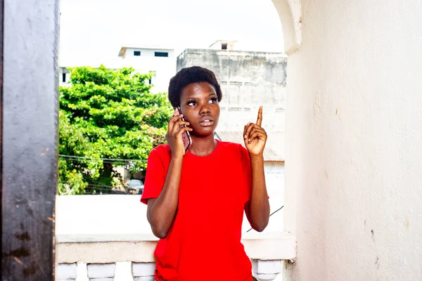 家のバルコニーで携帯電話で話している赤いTシャツを着た美しい少女 — ストック写真