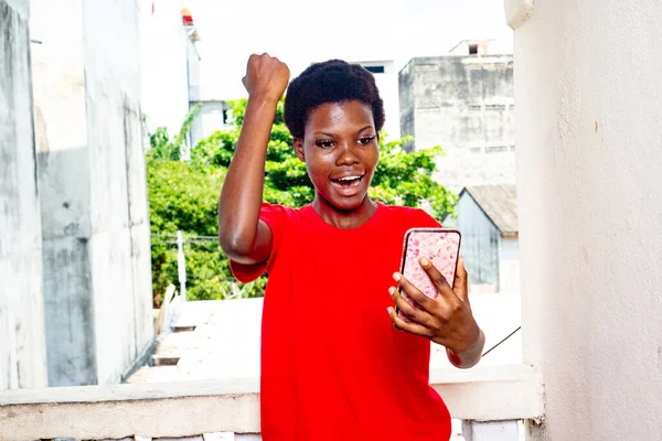 赤いTシャツを着た若い美少女が自宅で携帯電話を使って腕を上げて勝利のジェスチャーを作り — ストック写真