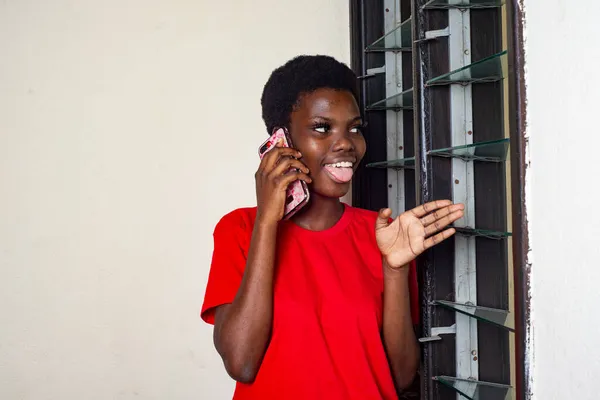 若いです美しい女の子です赤いTシャツに立って自宅で話している彼女の舌を突き出す窓の近くの携帯電話 — ストック写真