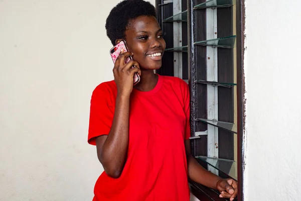 窓の外を見ながら携帯電話で話している家に立っている赤いTシャツの若い美しい女の子は微笑んだ — ストック写真