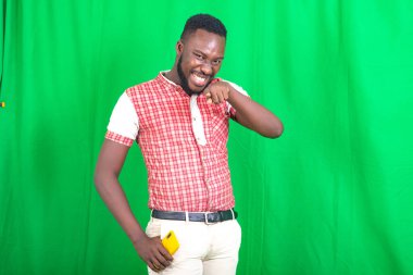 Tişörtlü, yeşil arka planda duran yakışıklı, sakallı bir adam kamerayı işaret ederek gülüyor..