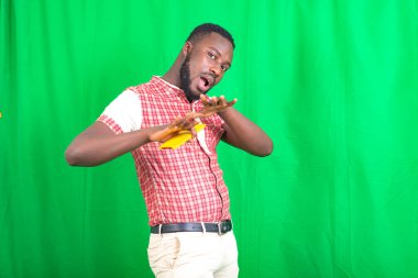 Yakışıklı, sakallı bir adam. Arka planda duruyor. Elleriyle surat yapıyor..