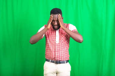 Yeşil arka planda duran ve baş ağrısı hisseden, tişörtlü, yakışıklı, sakallı bir adam..