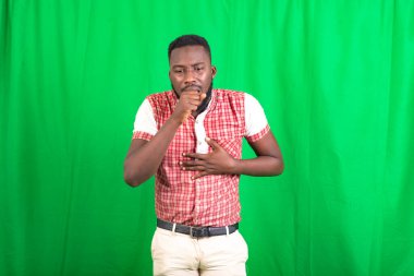 Tişörtlü, yeşil arka planda öksüren yakışıklı, sakallı bir adam..