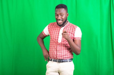 Tişörtlü, yeşil arka planda duran yakışıklı genç sakallı bir adam parmağını kameraya doğrultup gülümsüyor..