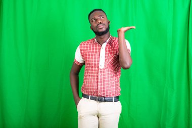 Tişörtlü, yeşil arka planda duran yakışıklı, sakallı bir adam. Avucunu düşünceli bir havayla sunuyor..