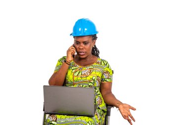 Güvenlik kaskı takan genç ve güzel bir bayan mühendis dizüstü bilgisayarla koltukta oturuyor ve gülümserken cep telefonuyla konuşuyor..