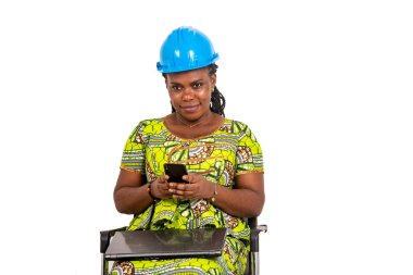 Güvenlik kaskı takan genç ve güzel bir bayan mühendis sandalyede oturuyor dizüstü bilgisayarlı ve cep telefonuyla sohbet ediyor..