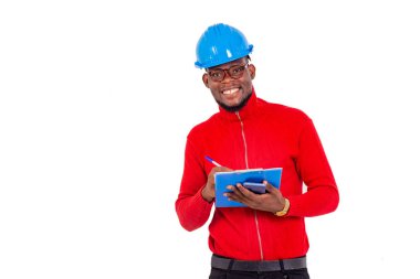 Genç bir mühendis, arka planda beyaz bir şapkayla kameraya bakıp gülümserken not defterine yazı yazıyor..