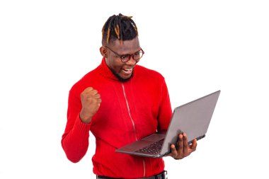 Kırmızı kazak giyen genç adam dizüstü bilgisayar kullanıyor ve gülümserken kolunu kaldırarak zafer işareti yapıyor..