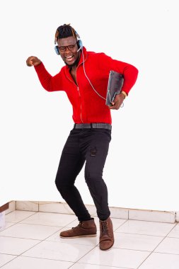 Beyaz arka planda duran, kulaklık ve dizüstü bilgisayarla gülerek müzik dinleyen genç bir öğrenci..