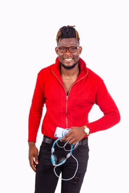 Gözlüklü genç bir adam beyaz arka planda duruyor elinde cep telefonu ve kulaklıkla kameraya bakıp gülümsüyor..