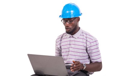 Güvenlik kaskı takan genç mühendis sandalyede oturuyor ve dizüstü bilgisayarla çalışıyor..