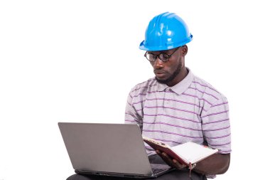 Genç mühendis kasketli, sandalyede oturmuş dizüstü bilgisayarla notları kontrol ediyor..