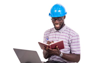 Genç mühendis kasketli sandalyede oturuyor ve dizüstü bilgisayarla not alıyor..