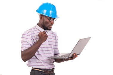 Genç mühendis mavi güvenlik kaskı takıyor, dizüstü bilgisayarı tutuyor ve yükseltilmiş koluyla zafer hareketi yapıyor..