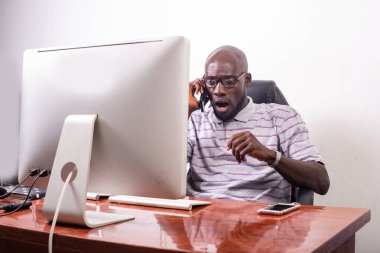 Genç şaşkın işadamı masanın başında dizüstü bilgisayara bakıyor ve cep telefonuyla konuşuyor..