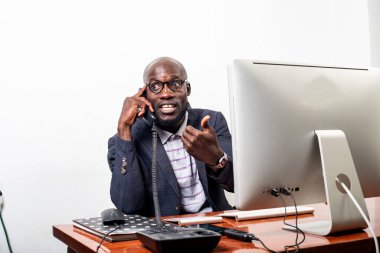 Genç iş adamı dizüstü bilgisayar karşısında oturuyor ve gülümserken cep telefonuyla konuşuyor..